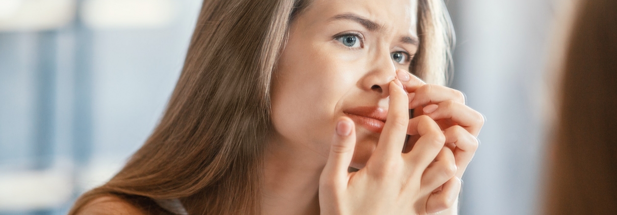 Displeased young woman squeezing acne on her nose while looking in mirror indoors