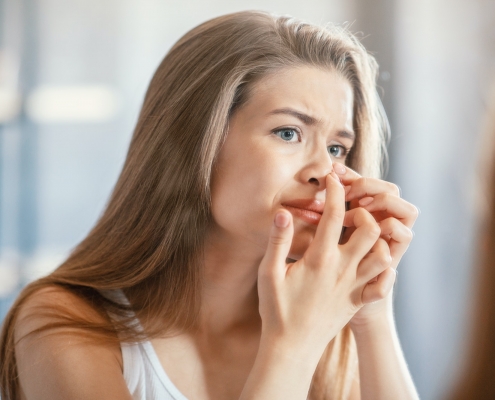Displeased young woman squeezing acne on her nose while looking in mirror indoors